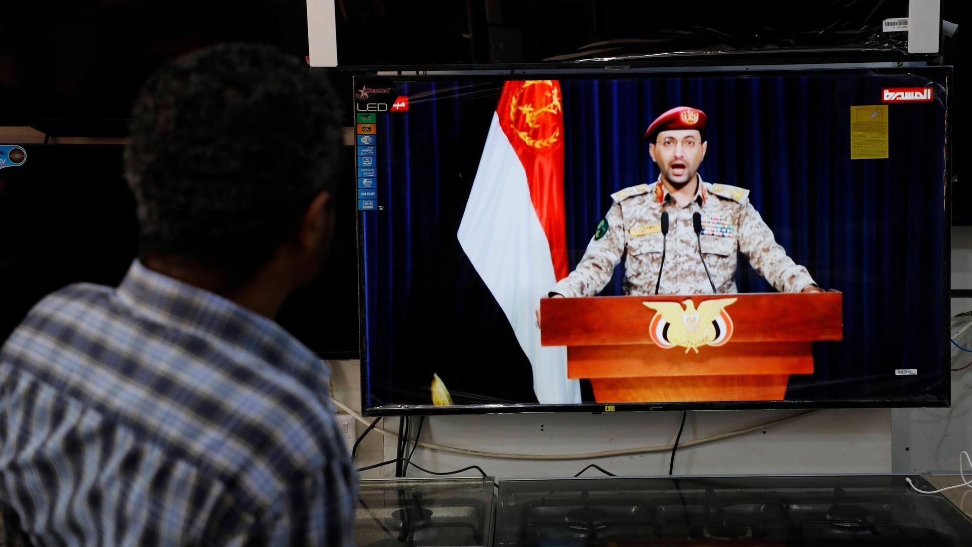 Ein Sprecher der Huthi-Rebellen gibt im Fernsehen ein Statement zum Angriff auf das Schiff ab. Davor steht ein Mann und hört zu.