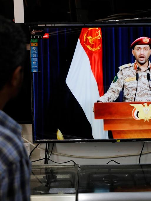 Ein Sprecher der Huthi-Rebellen gibt im Fernsehen ein Statement zum Angriff auf das Schiff ab. Davor steht ein Mann und hört zu.