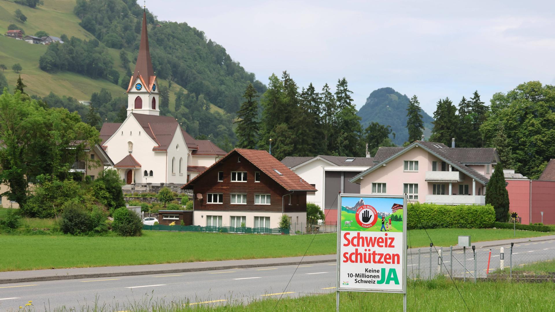 Das Plakat "Schweiz schützen. Keine 10 Millionen Schweiz" steht in einer Berglandschaft mit einem Schweizer Dorf im Hintergrund. Das Plakat "Schweiz schützen. Keine 10 Millionen Schweiz" steht in einer Berglandschaft mit einem Schweizer Dorf im Hintergrund.