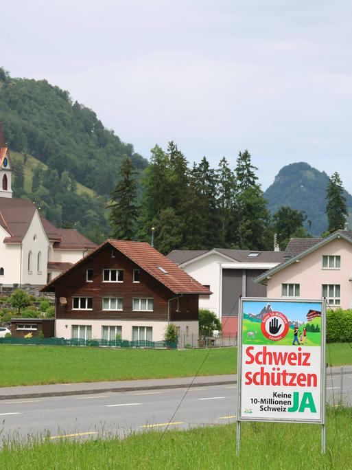 Das Plakat "Schweiz schützen. Keine 10 Millionen Schweiz" steht in einer Berglandschaft mit einem Schweizer Dorf im Hintergrund. Das Plakat "Schweiz schützen. Keine 10 Millionen Schweiz" steht in einer Berglandschaft mit einem Schweizer Dorf im Hintergrund.