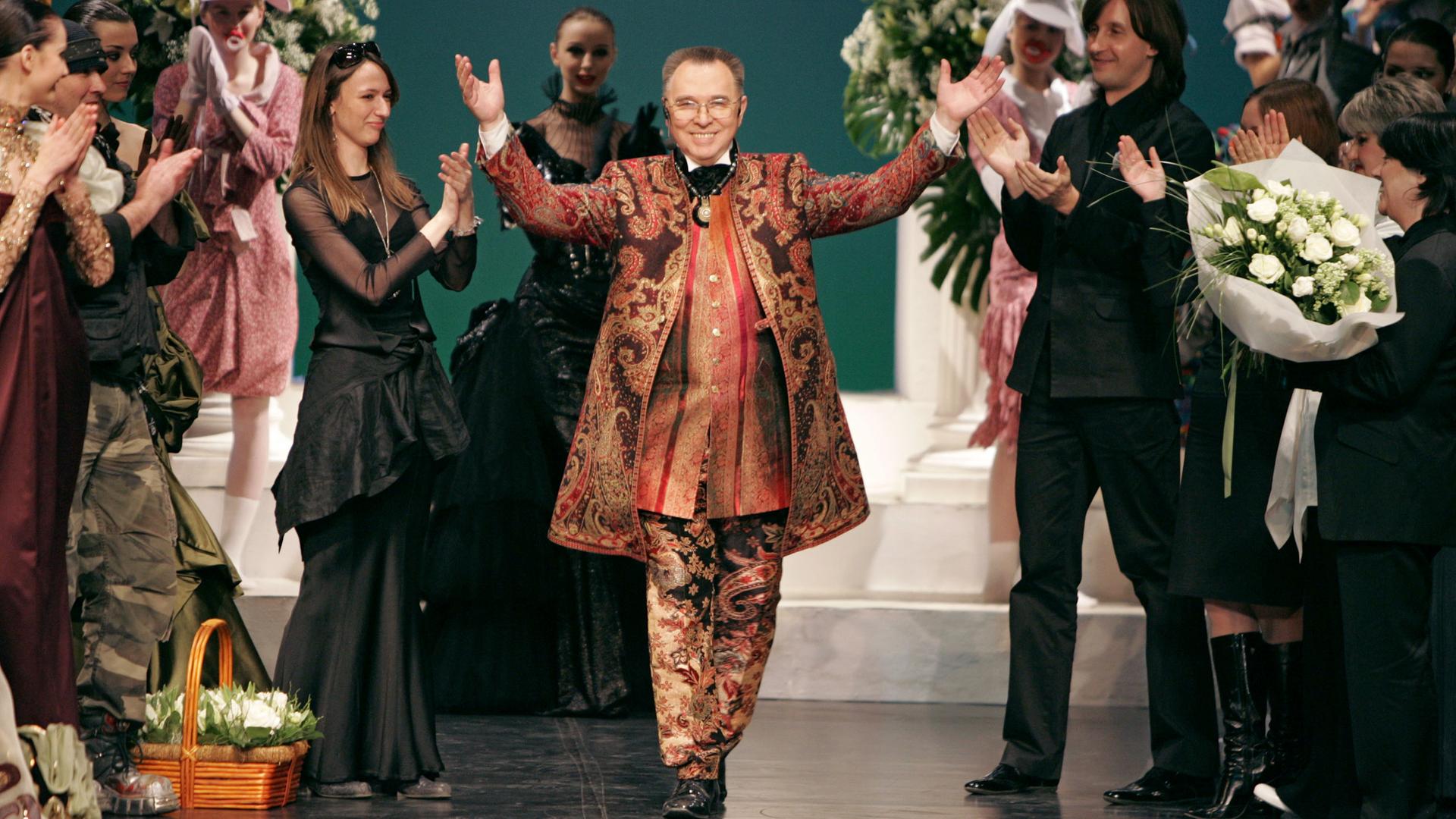Der russische Modedesigner Slava Zaitsev läuft mit erhobenen Händen über eine Bühne. Er trägt Brille und einen Mantel, ein Hemd und eine Hose mit floralem Design. Neben ihm stehen applaudierende Models.