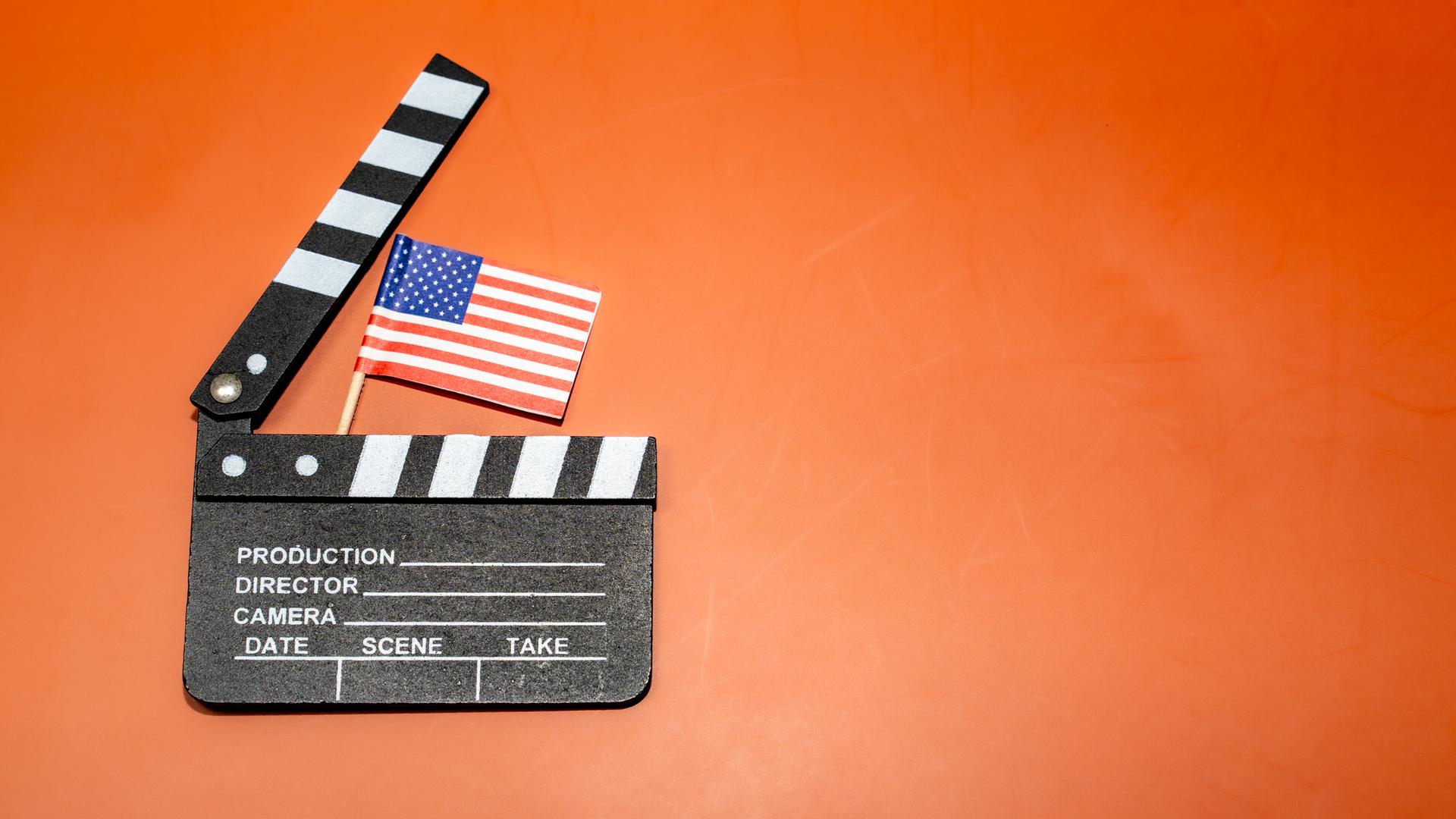Auf einem leuchtend orangefarbenen Hintergrund ist eine Filmklappe mit einer amerikanischen Flagge zu sehen, die für das Filmemachen und patriotische Themen steht.