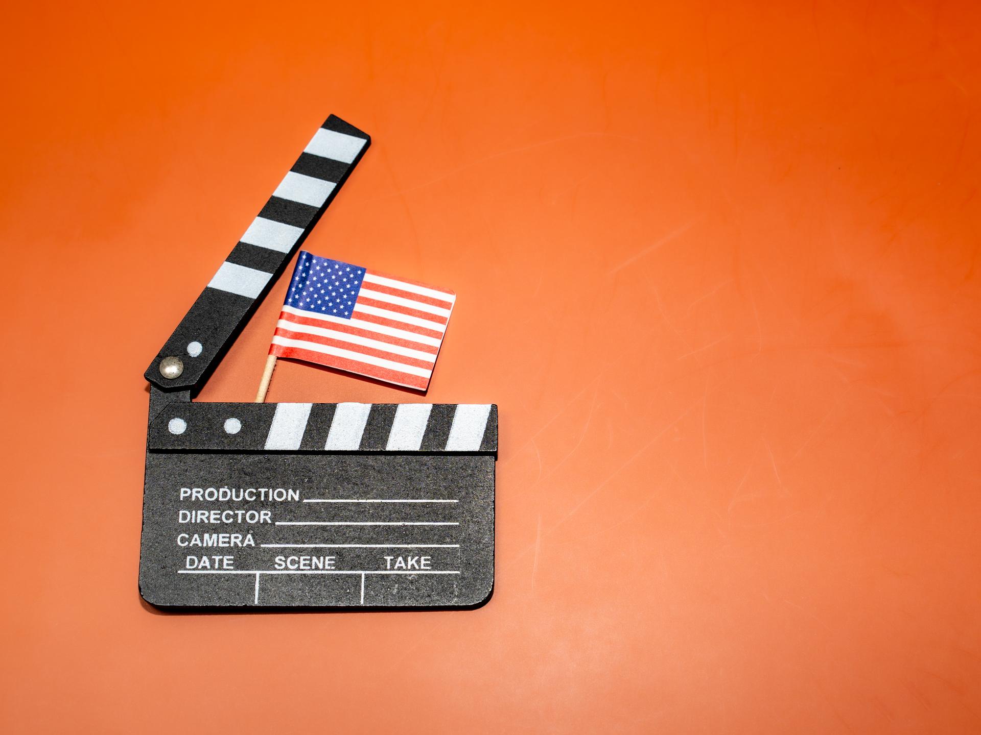 Auf einem leuchtend orangefarbenen Hintergrund ist eine Filmklappe mit einer amerikanischen Flagge zu sehen, die für das Filmemachen und patriotische Themen steht.