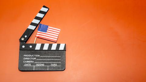 Auf einem leuchtend orangefarbenen Hintergrund ist eine Filmklappe mit einer amerikanischen Flagge zu sehen, die für das Filmemachen und patriotische Themen steht.