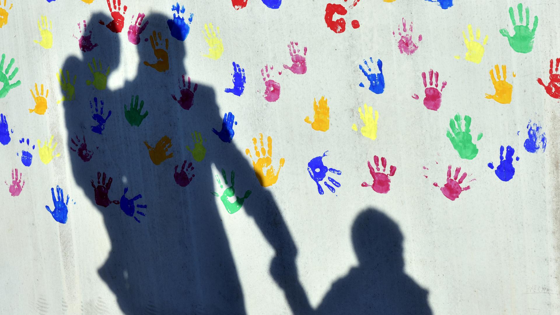 Ein Mann mit einem Kind auf dem Arm und einem an der Hand wirft einen Schatten auf eine mit bunten Handabdrücken bemalte Wand einer Kindertagesstätte.