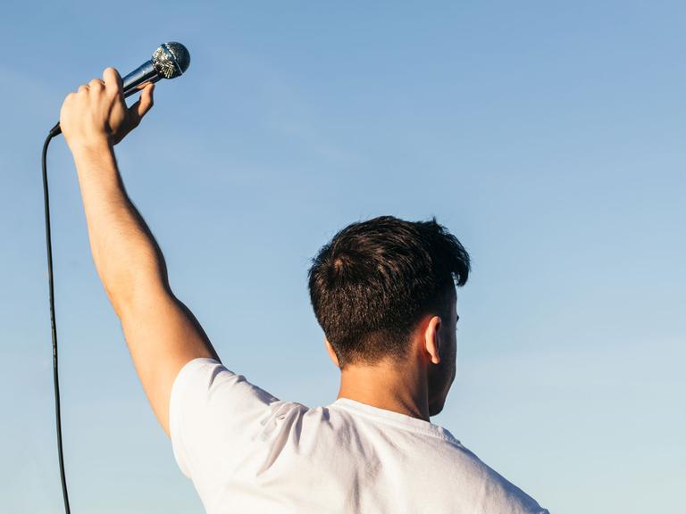Zu sehen: Ein junger Mann hält ein Mikrofon in die Luft