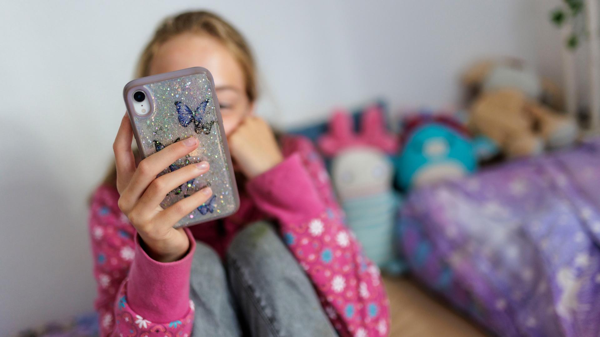 Ein Mädchen sitzt in ihrem Bett und schaut auf ihr Mobiltelefon. Ihr Gesicht ist nicht zu sehen. Ein Mädchen sitzt in ihrem Bett und schaut auf ihr Mobiltelefon. Ihr Gesicht ist nicht zu sehen.