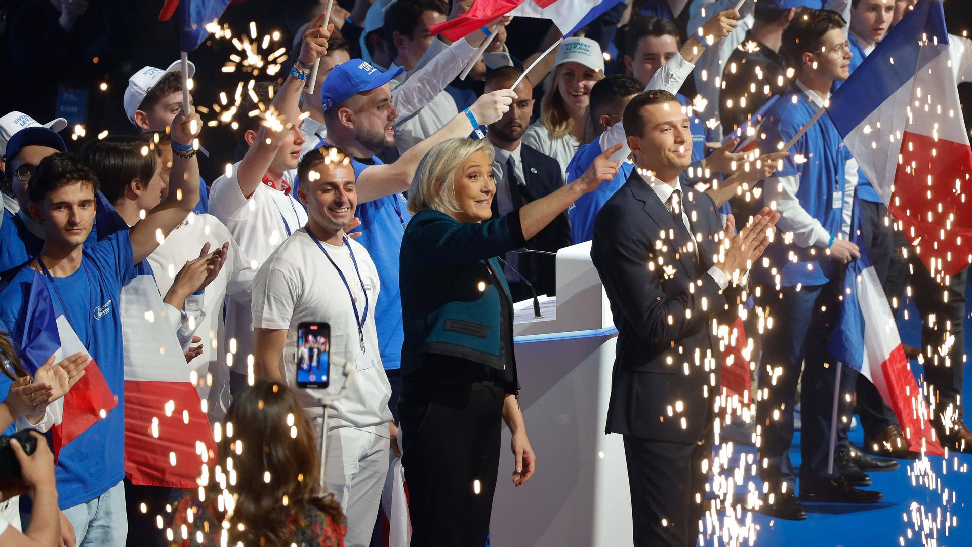 Marine Le Pen und ihr Parteichef Bardella vom Rassemblement National stehen auf einer Bühne, umringt von Anhängern, und feiern.