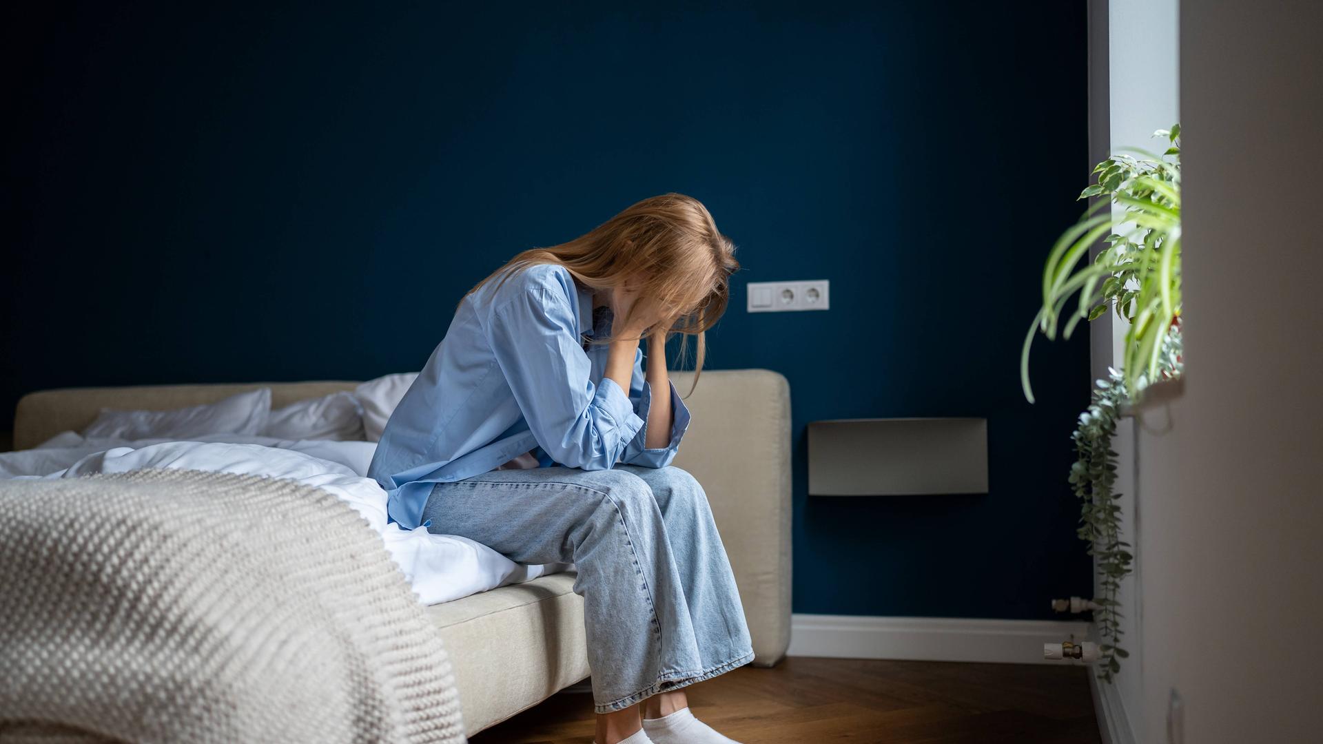 Eine junge Frau sitzt auf dem Bett im Schlafzimmer und stützt ihr Gesicht in die Hände,.