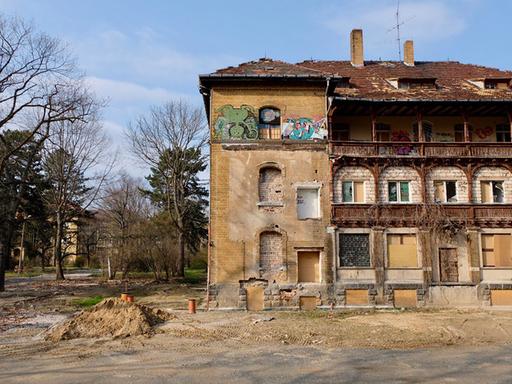 Blick auf ein heruntergekommen aussehendes Gebäude der ehemaligen Psychiatrie in Leipzig-Dösen. Daneben liegt ein kleiner Erdhügel und im Bolden stecken einige Rohre. Im Hintergrund ist in dem parkähnlichen Gelände ein weiteres Gebäude zu sehen. 