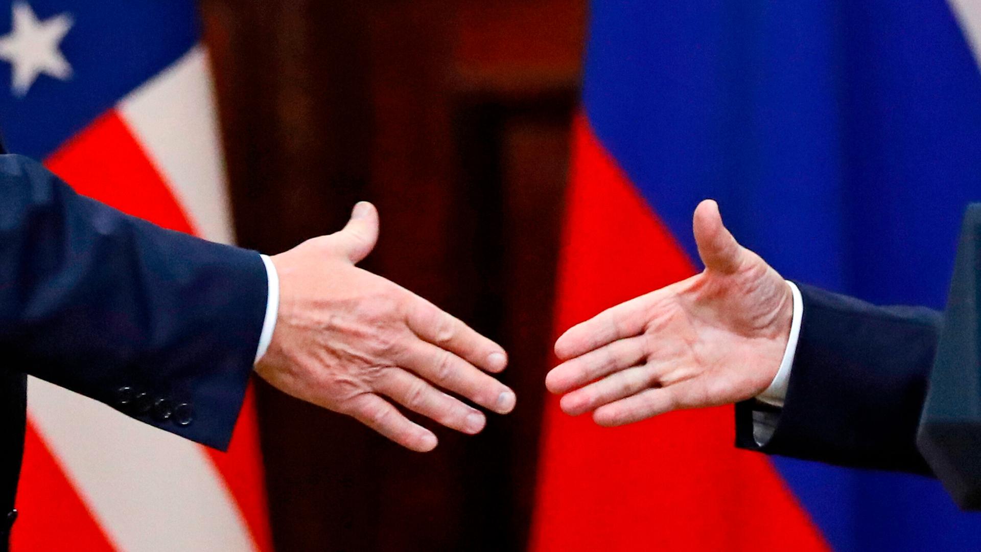 US-Präsident Donald Trump  und Russlands Machthaber Wladimir Putin geben sich die Hände: Großaufnahme der Hände, im Hintergrund die russische und US-amerikanische Flagge.