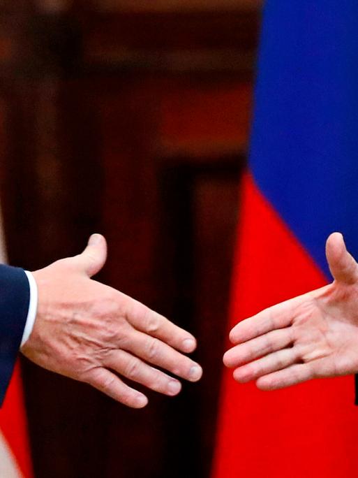 US-Präsident Donald Trump  und Russlands Machthaber Wladimir Putin geben sich die Hände: Großaufnahme der Hände, im Hintergrund die russische und US-amerikanische Flagge.