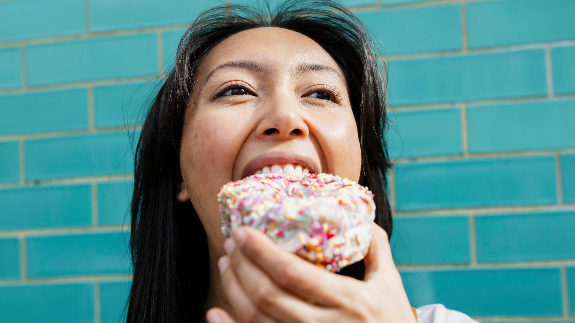 Lächelnde Frau isst einen mit Zuckerstreuseln gesprinkelten Donut vor einer türkisen Wand