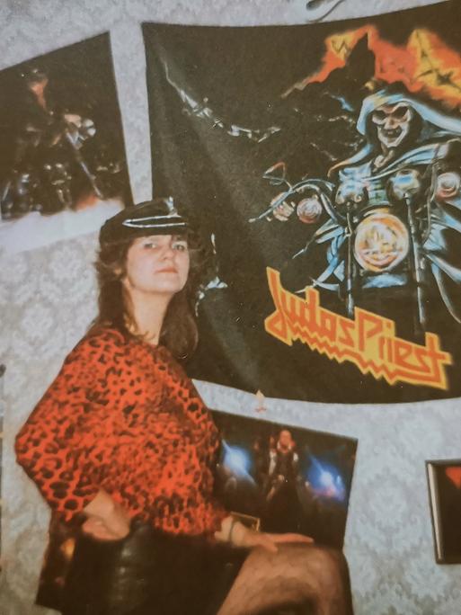 Eine Frau mit lederner Mütze und in rotem Oberteil mit Leopardenmuster steht vor einer Wand mit ornamentaler Tapete in Beigetönen und mehreren daran befestigten Postern der Band Judas Priest.