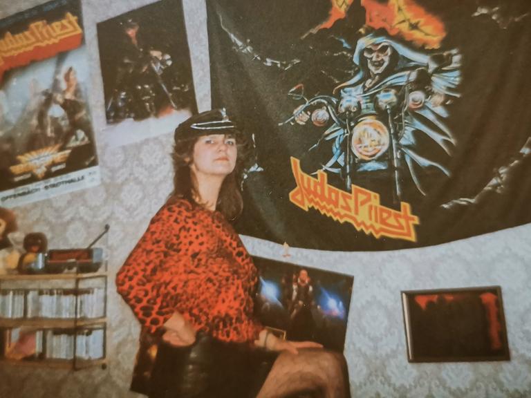Eine Frau mit lederner Mütze und in rotem Oberteil mit Leopardenmuster steht vor einer Wand mit ornamentaler Tapete in Beigetönen und mehreren daran befestigten Postern der Band Judas Priest.