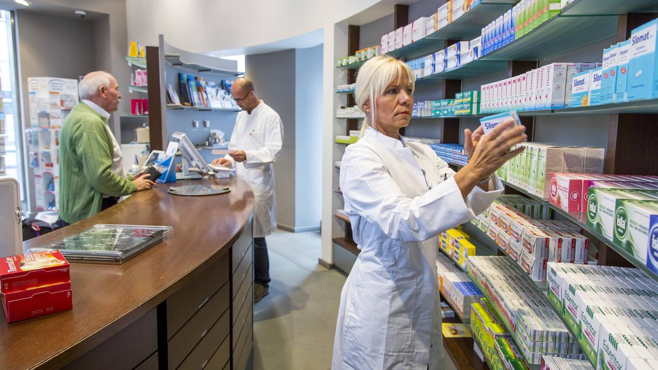 Eine Apothekerin steht hinter einem Verkaufsthresen und greift eine Medikamentenpackung aus dem Regal, im Hintergrund bearbeitet ein Apotheker das Rezept eines älteren Herrn.