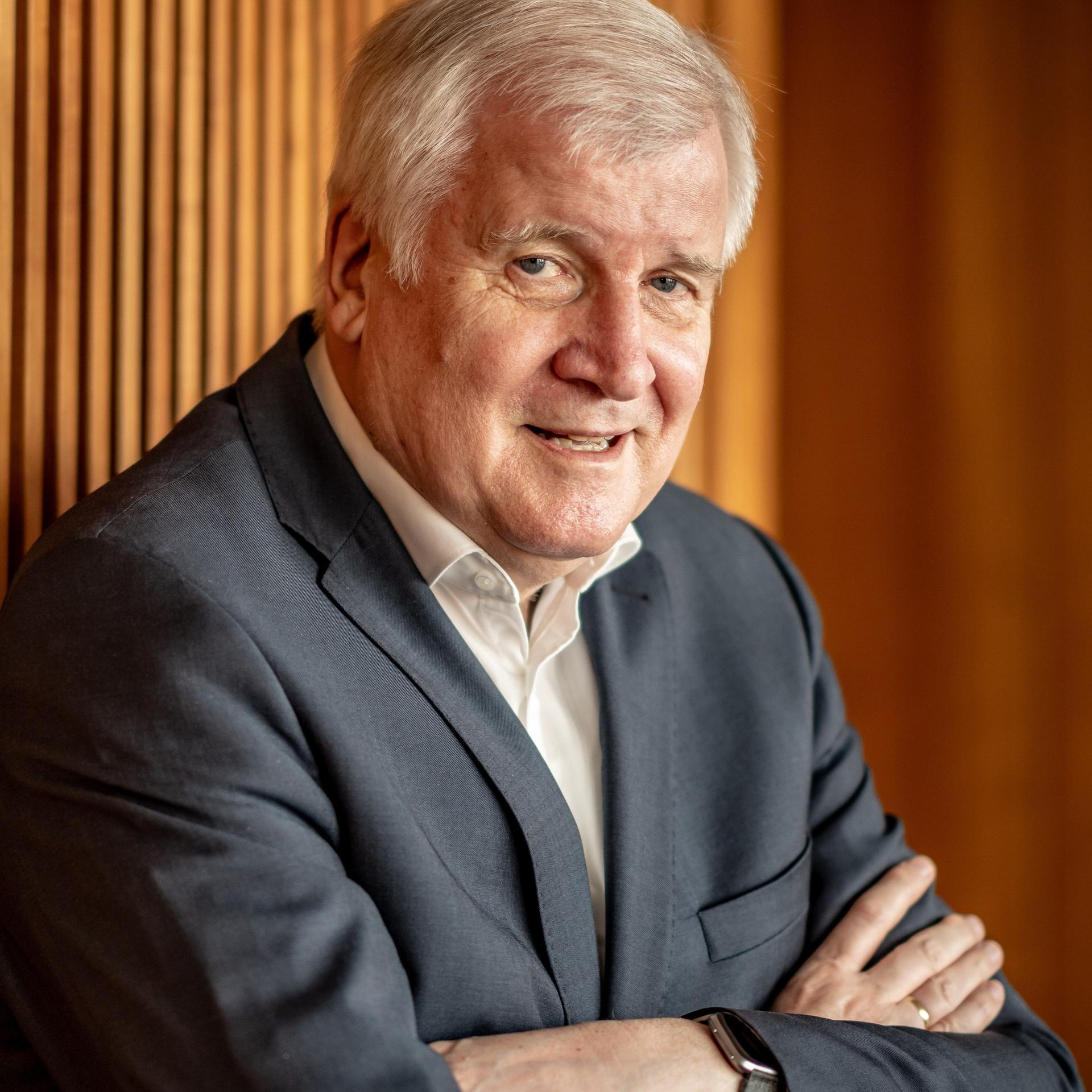 Horst Seehofer lehnt mit verschränkten Armen an einer Wand mit Holzpaneelen und lächelt in die Kamera.