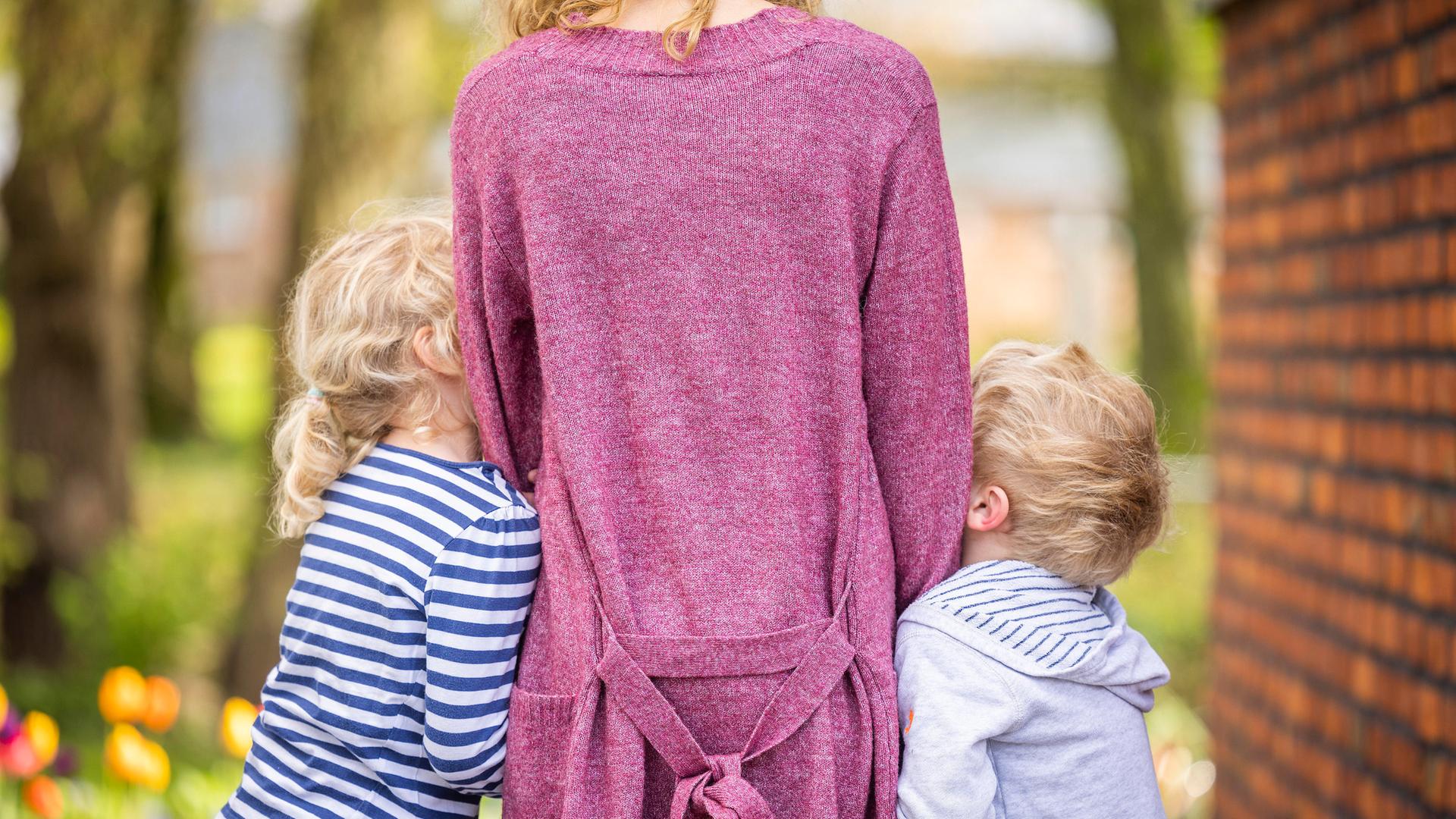 Eine stehende Frau ist von hinten zu sehen – mit zwei Kindern, die sich rechts und links an sie schmiegen.