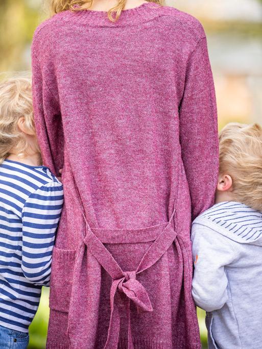 Eine stehende Frau ist von hinten zu sehen – mit zwei Kindern, die sich rechts und links an sie schmiegen.