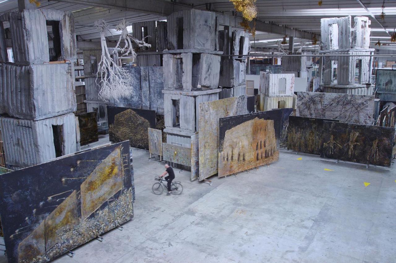 Der Künstler Anselm Kiefer fährt auf einem Fahrrad über sein Ateliergelände. Es sind großformatige Leinwände zu sehen.