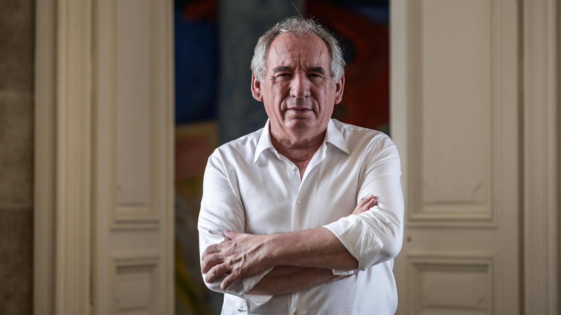 François Bayrou. Ein älterer Herr mit grauem Haar und im weißen Hemd steht in einem Raum und schaut direkt in die Kamera.