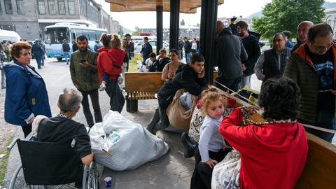 Menschen, unter ihnen Kinder und ein älterer Mann im Rollstuhl, stehen an einem Unterstand. Im Hintergrund ein Reisebus.