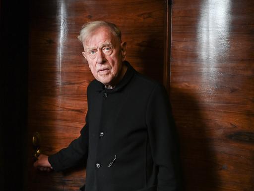 Claus Peymann steht vor einer Holzvertäfelung und blick ernst in die Kamera