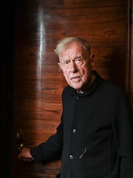 Claus Peymann steht vor einer Holzvertäfelung und blick ernst in die Kamera