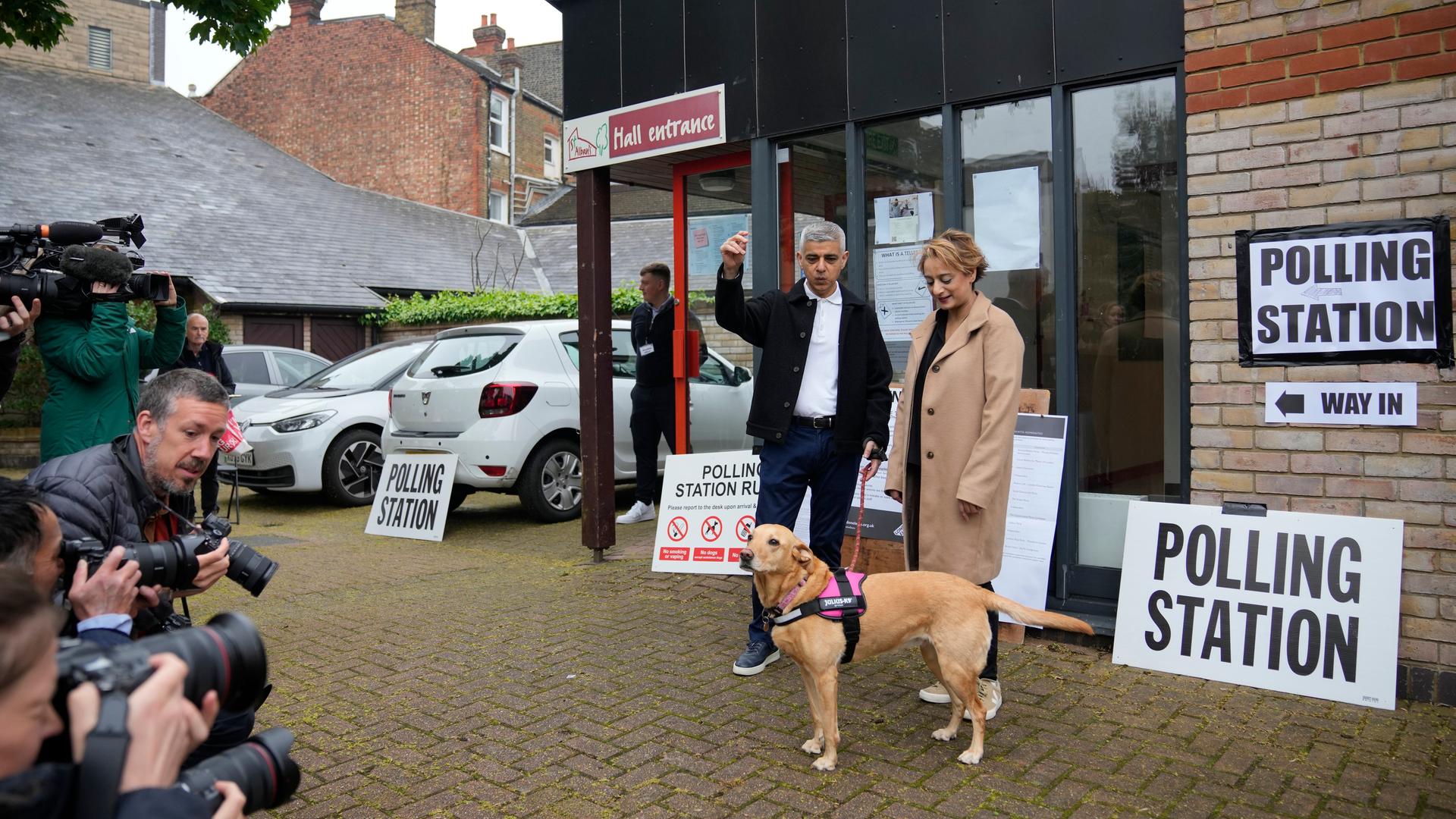 Die beiden stehen mit ihrem braunen Hund vor der "Polling Station" und werden von Fotografen fotografiert. Khan hebt die Hand nach oben. 