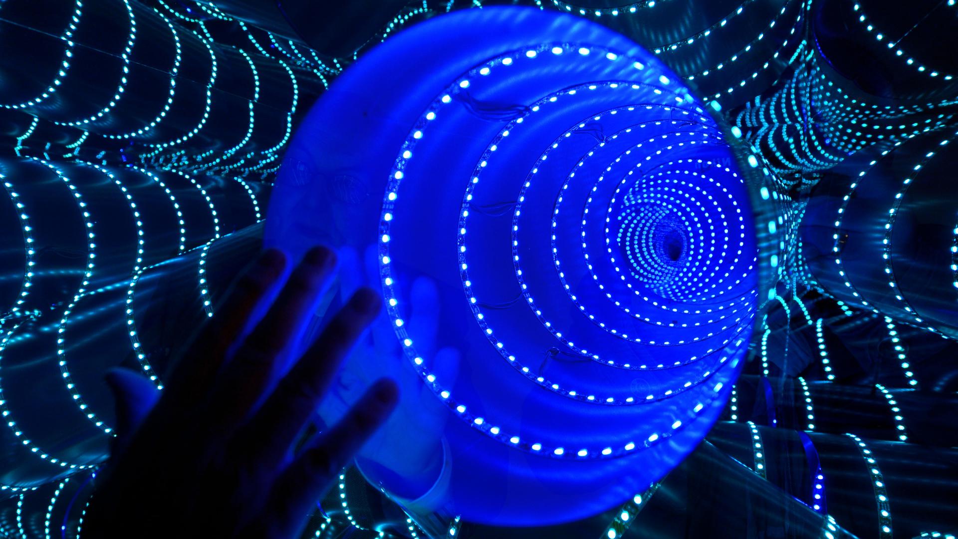Schattenriss einer Hand vor dem Licht-Kunstwerk "triple tube" des Künstlers Hans Kotter. Ein blauer Lichtunnel in der Mitte ist von Lichtgirlanden umgeben. Schattenriss einer Hand vor dem Licht-Kunstwerk "triple tube" des Künstlers Hans Kotter. Ein blauer Lichtunnel in der Mitte ist von Lichtgirlanden umgeben.