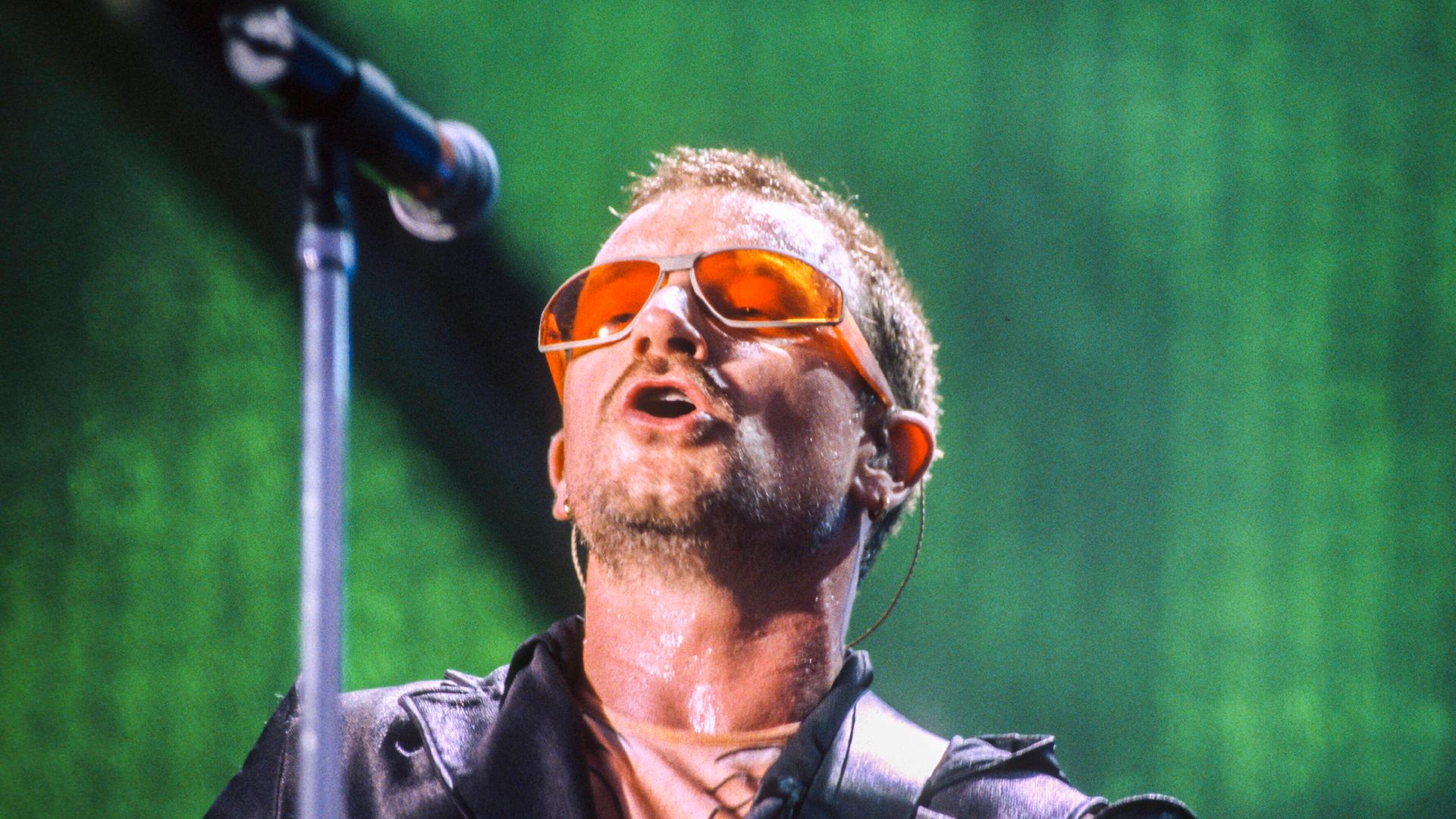 Nahaufnahme eines Mannes mit einer Kurzhaarfrisur und Dreitagebart singend an einem Mikrofon mit eine Brille mit orangefarbenen Gläsern und schwarzer Jacke. Nahaufnahme eines Mannes mit einer Kurzhaarfrisur und Dreitagebart singend an einem Mikrofon mit eine Brille mit orangefarbenen Gläsern und schwarzer Jacke.