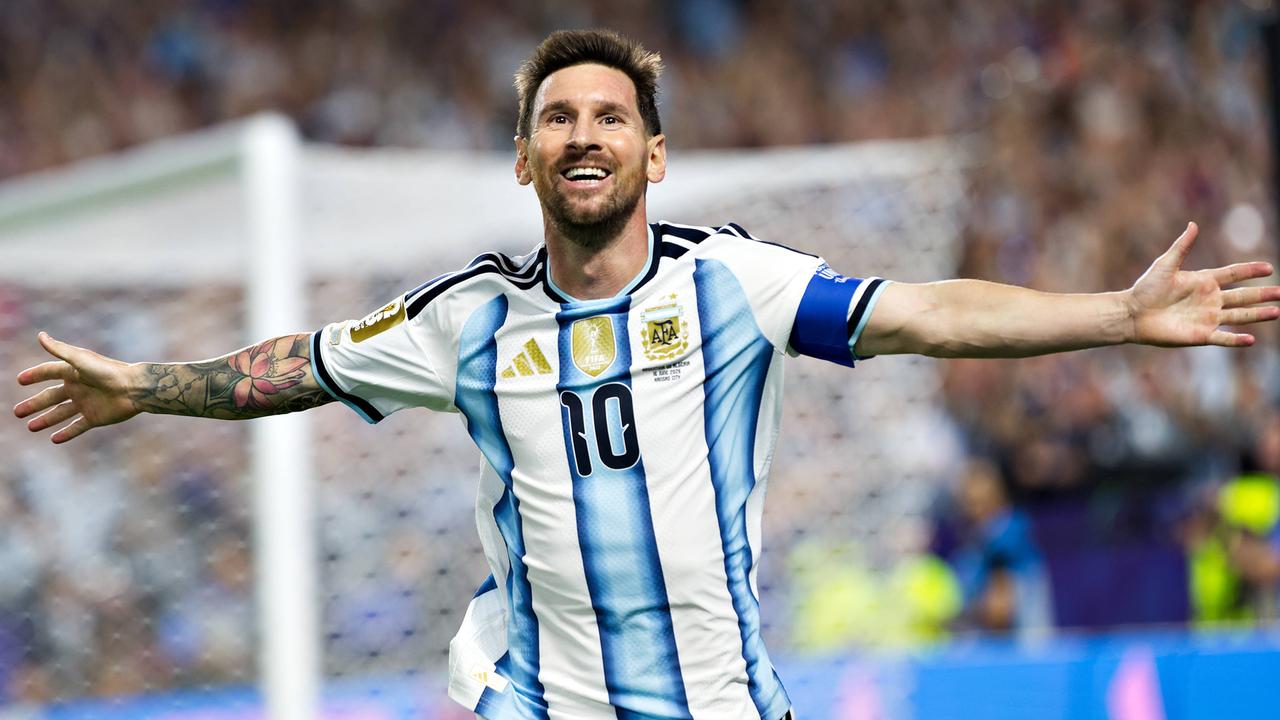 Lionel Messi läuft im blau-weiß gestreiften Trikot mit ausgebreiteten Armen über den Fußballrasen, im Hintergrund sind verschwommen ein Tor und Zuschauer zu sehen.