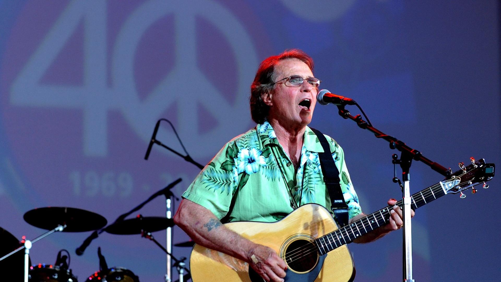 Der Country-Musiker Joe McDonald beim Bethel Woods Music Festival 2009 mit seiner Gitarre in der Hand, hinter ihm eine 40 mit einem Peace-Zeichen als Null