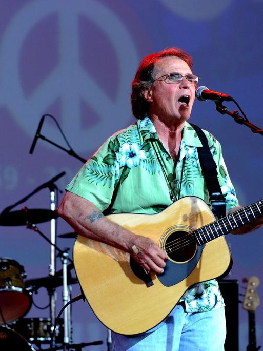 Der Country-Musiker Joe McDonald beim Bethel Woods Music Festival 2009 mit seiner Gitarre in der Hand, hinter ihm eine 40 mit einem Peace-Zeichen als Null