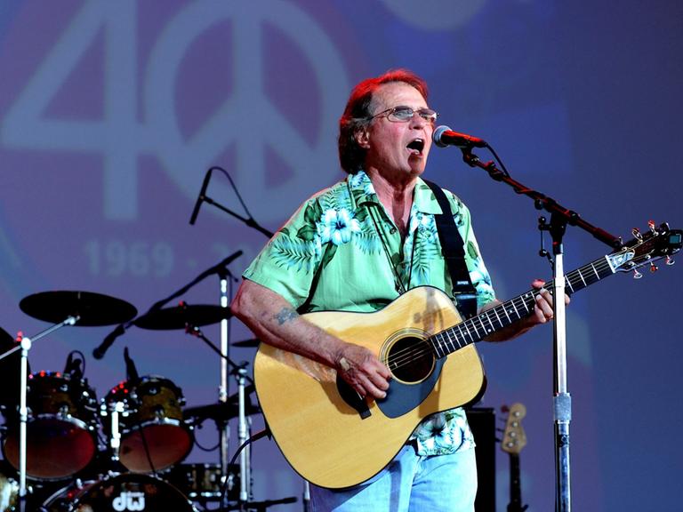 Der Country-Musiker Joe McDonald beim Bethel Woods Music Festival 2009 mit seiner Gitarre in der Hand, hinter ihm eine 40 mit einem Peace-Zeichen als Null