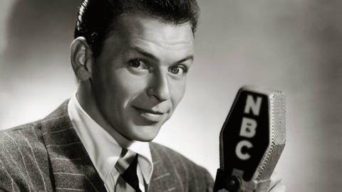 Schwarzweiß-Porträt von Frank Sinatra, der in die Kamera lächelt und ein altmodisches Mikrofon in der Hand hält.