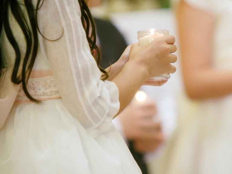 Junge Mädchen, deren Gesichter nicht zu erkennen sind, halten eine Kerze im Glas in der Hand. Sie tragen festliche weiße Kleidung.