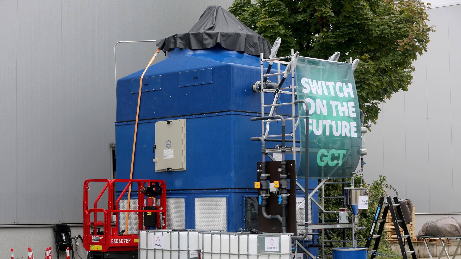 Das Bild zeight einen großen blauen Container mit Rohren vor der Wand einer weißen Halle. Auf einem Schild davor steht "Switch on the Future". Das Bild zeight einen großen blauen Container mit Rohren vor der Wand einer weißen Halle. Auf einem Schild davor steht "Switch on the Future".