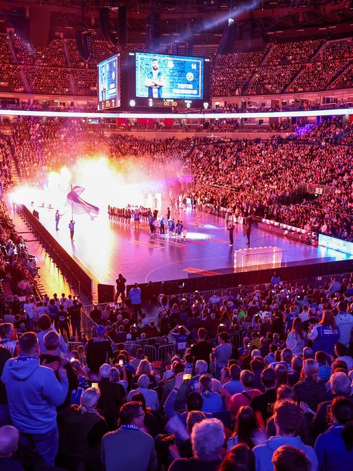 Blick in die abgedunkelte KölnArena, eine sehr große Sporthalle, die Ränge voller Zuschauer, in der Mitte das Handballfeld, auf dem mit Lichteffekten die Mannschaften einlaufen.