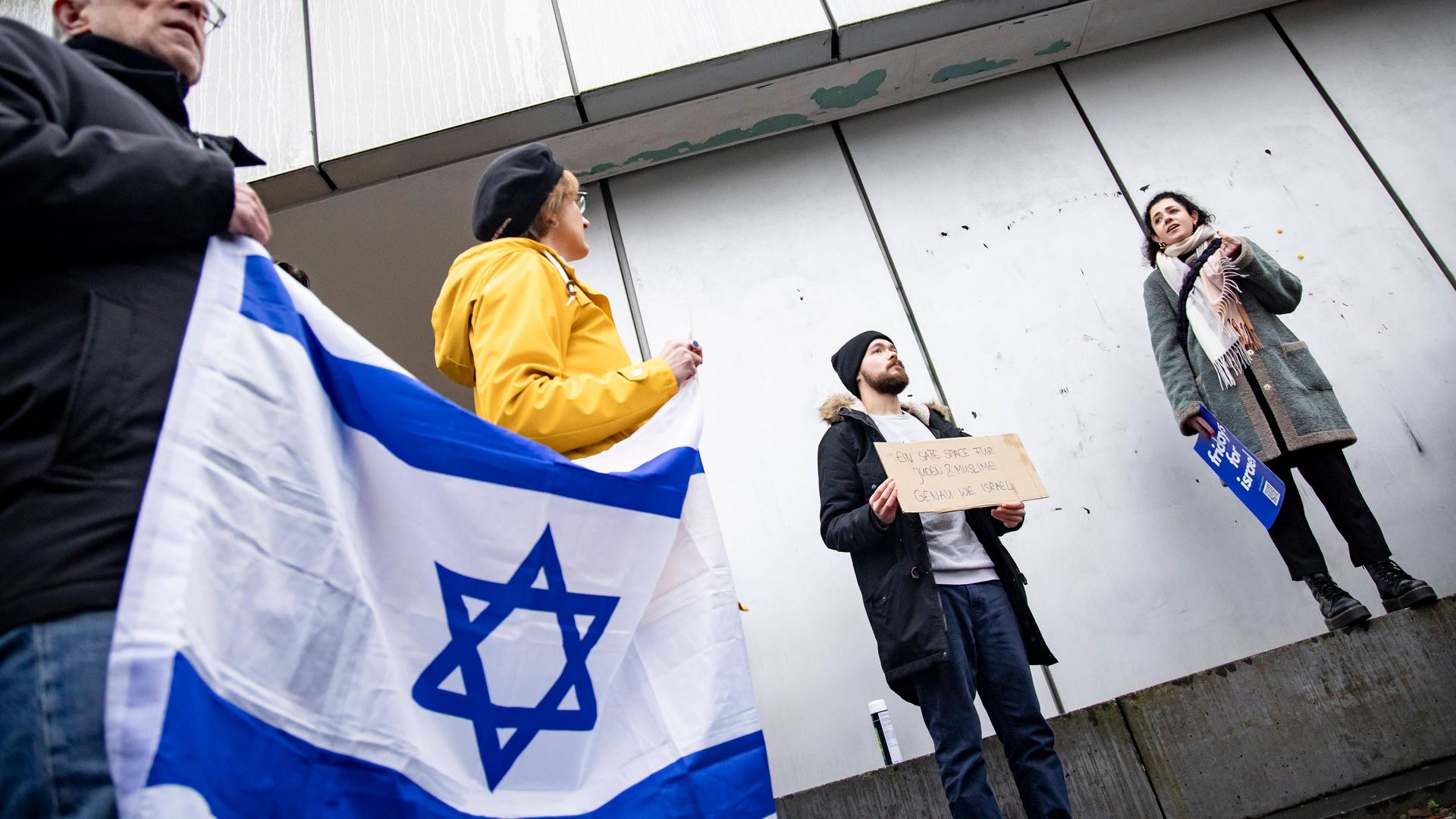Mehrere Menschen stehen während am Rande einer Demonstration, halten eine Rede und zeigen eine Israel-Flagge.