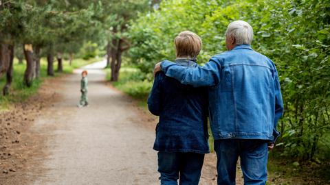 Ein junges Rentnerpaar spaziert durch einen Wald. Der Mann hat den Arm um die Frau gelegt, vorn hinaus läuft ein Kind. 