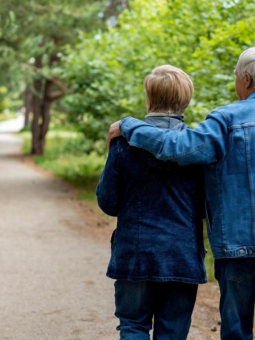 Ein junges Rentnerpaar spaziert durch einen Wald. Der Mann hat den Arm um die Frau gelegt, vorn hinaus läuft ein Kind. 