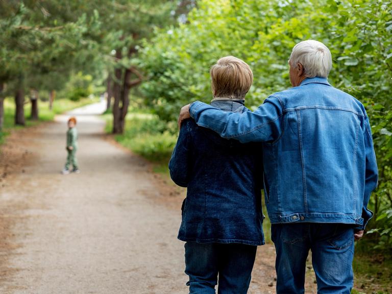 Ein junges Rentnerpaar spaziert durch einen Wald. Der Mann hat den Arm um die Frau gelegt, vorn hinaus läuft ein Kind. 