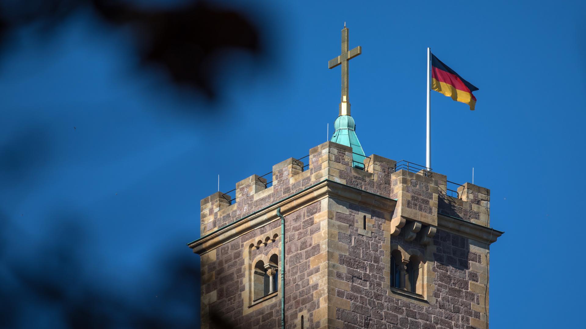 Die Sonne scheint auf die Deutschlandfahne auf der Wartburg in Eisenach (Thüringen). Daneben ist ein Turmkreuz zu sehen.