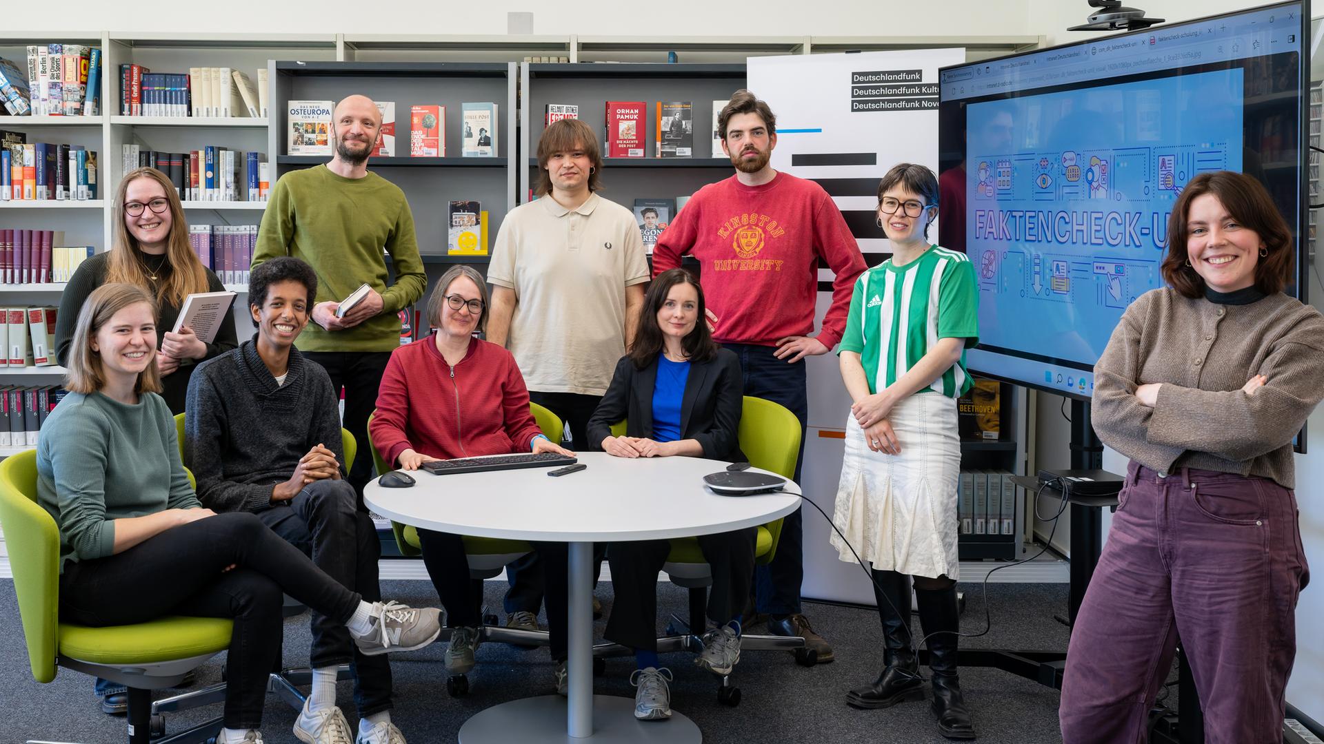 Ein Team aus mehreren Frauen und Männern stehen und sitzen um einen runden Tisch herum vor einem Bücherregal Ein Team aus mehreren Frauen und Männern stehen und sitzen um einen runden Tisch herum vor einem Bücherregal