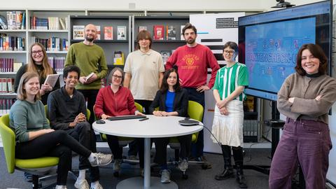 Ein Team aus mehreren Frauen und Männern stehen und sitzen um einen runden Tisch herum vor einem Bücherregal