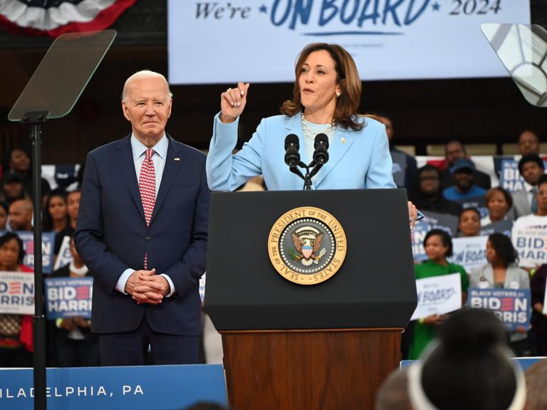 Archivbild: Kamala Harris steht bei einer Wahlkampfveranstaltung am Rednerpult und spricht zum Publikum, links hinter ihr steht Joe Biden. Im Hintergrund sind Unterstützer der Demokraten mit Wahlkampfplakaten in den Händen zu sehen.