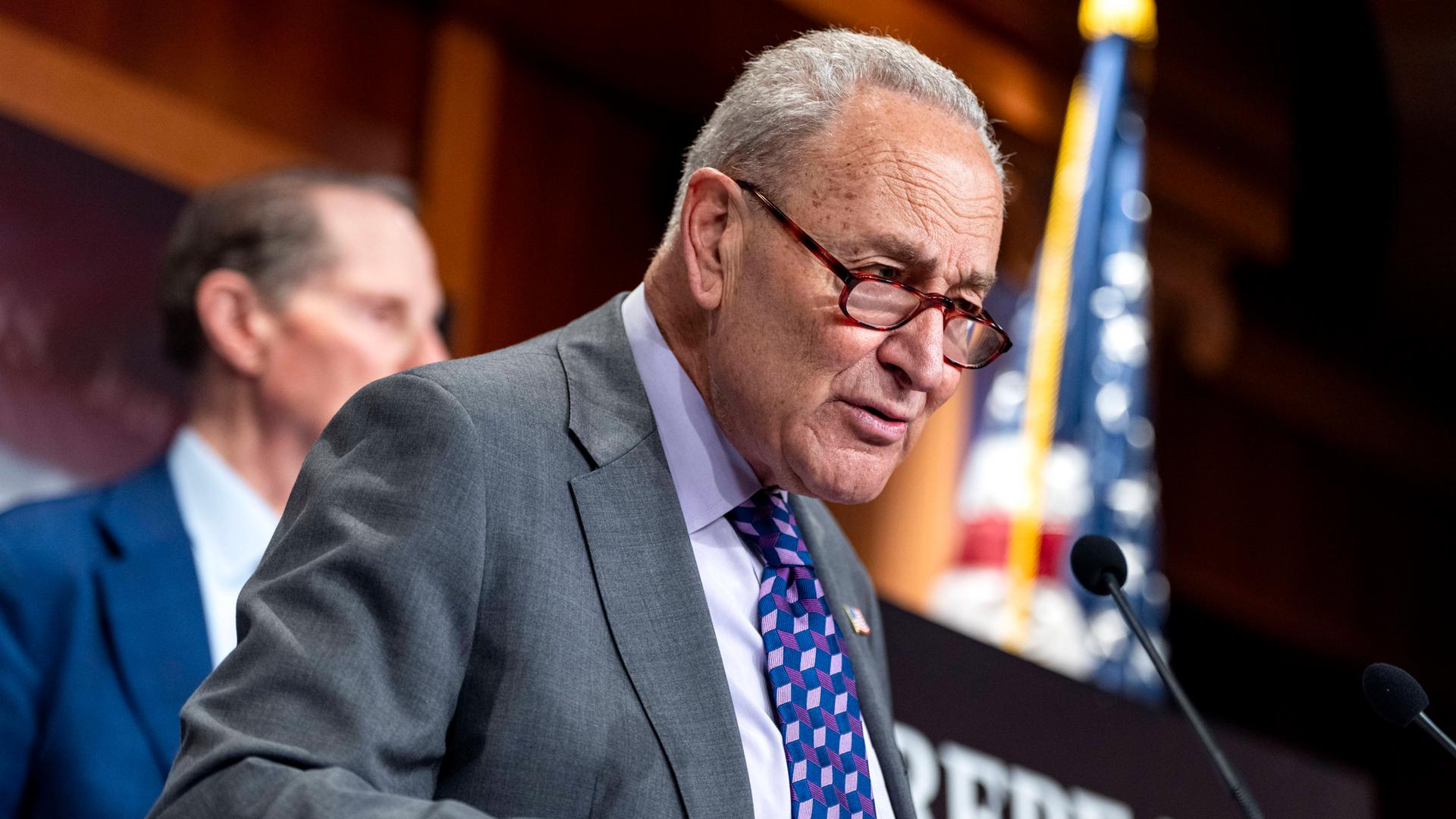 Chuck Schumer steht an einem Rednerpult und spricht in ein Mikrofon. Im Hintergrund ist eine US-amerikanische Flagge. Chuck Schumer steht an einem Rednerpult und spricht in ein Mikrofon. Im Hintergrund ist eine US-amerikanische Flagge.