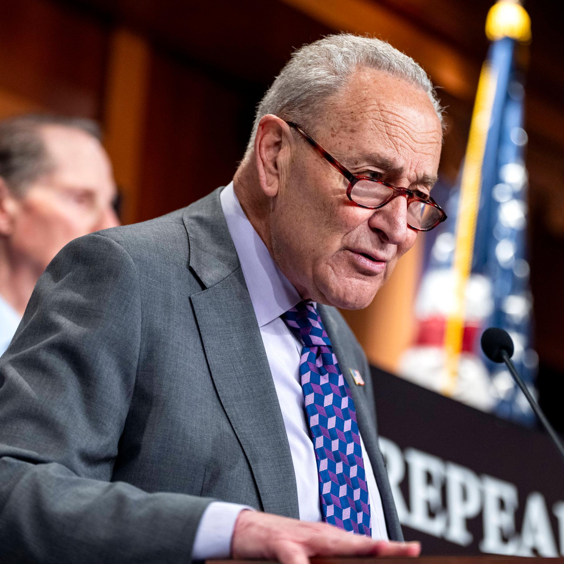 Schumer steht an einem Rednerpult und spricht in ein Mikrofon. Im Hintergrund ist eine US-amerikanische Flagge.