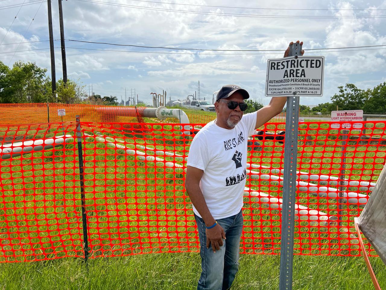 Ein Mann mit Käppi, Sonnenbrille und weißem T-Shirt steht an einem roten Zaun vor einem Schild auf dem "Restricted Area" steht.